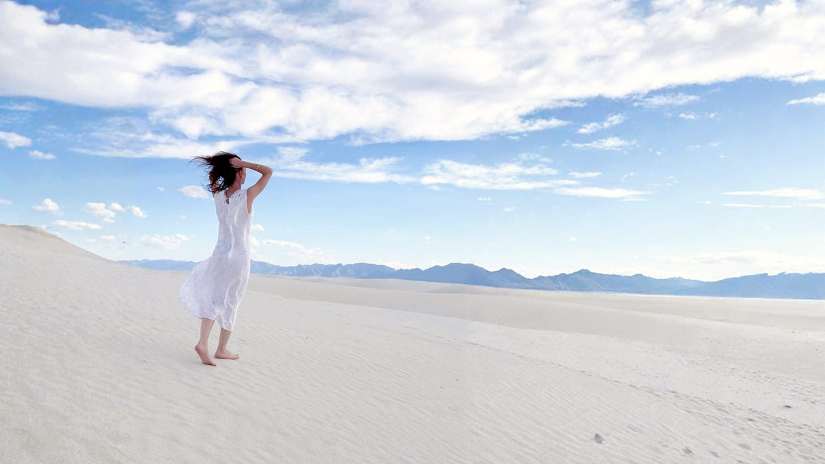 Kalmeon de dos, en robe blanche, les cheveux au vent sur des dunes de sable blanc sous un ciel bleu