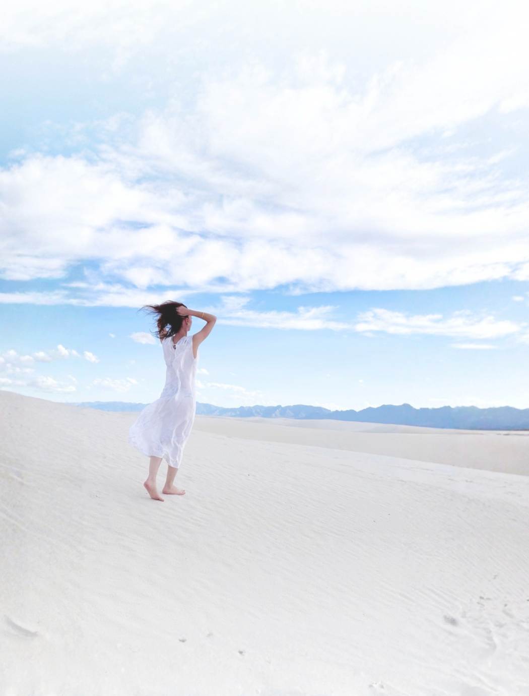 Kalmeon de dos, en robe blanche, les cheveux au vent sur des dunes de sable blanc sous un ciel bleu